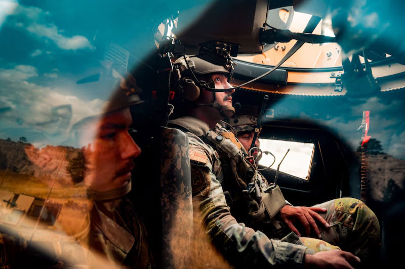 Three service members wearing combat gear sit inside the cockpit of a military vehicle..