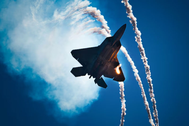 A military aircraft flies overhead against a blue sky leaving a trail of smoke..